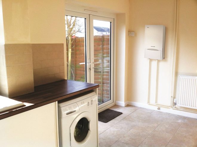 Photo showing a modern kitchen diner room in a domestic house, with white glossy cabinets, an upright automatic washing machine, a ceramic sink located in front of a window, beige wall tiles and a brown tiled floor. The floor tiles have a mottled pattern, which helps to disguise a dirty floor. Double glazed patio doors / French windows lead into the back garden, where there is a lawn, wooden larch-lap fence panels, various flower pots and plants, and a wood picnic table. A doormat can be seen by the concrete paving slabs.