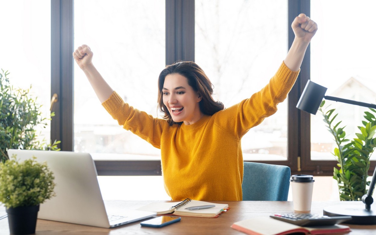 Vesela ženska sedi za računalnikom. Oblečena je v rumen pulover