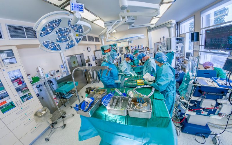 Fotografija prikazuje ekipo kirurgov med operacijo v moderni kirurški operacijski sobi. Okolje je opremljeno z napredno medicinsko opremo, sterilnimi instrumenti in monitorji, ki prikazujejo vitalne znake pacienta. Slika poudarja visoko stopnjo strokovnosti in tehnološko naprednost v sodobni medicini. Primerna je za članke o kirurgiji, zdravstveni oskrbi, medicinskih tehnologijah in bolnišničnih postopkih.