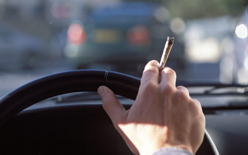 Fotografija prikazuje roko voznika, ki drži volan, medtem ko v drugi roki drži prižgano marihuano. Vozilo je v prometu, kar simbolizira vožnjo pod vplivom marihuane in opozarja na tveganja in nevarnosti, povezane s takšnim ravnanjem. Ta slika je pomembna za ozaveščanje o varnosti v prometu, odgovorni vožnji in zakonodaji glede vožnje pod vplivom drog.
