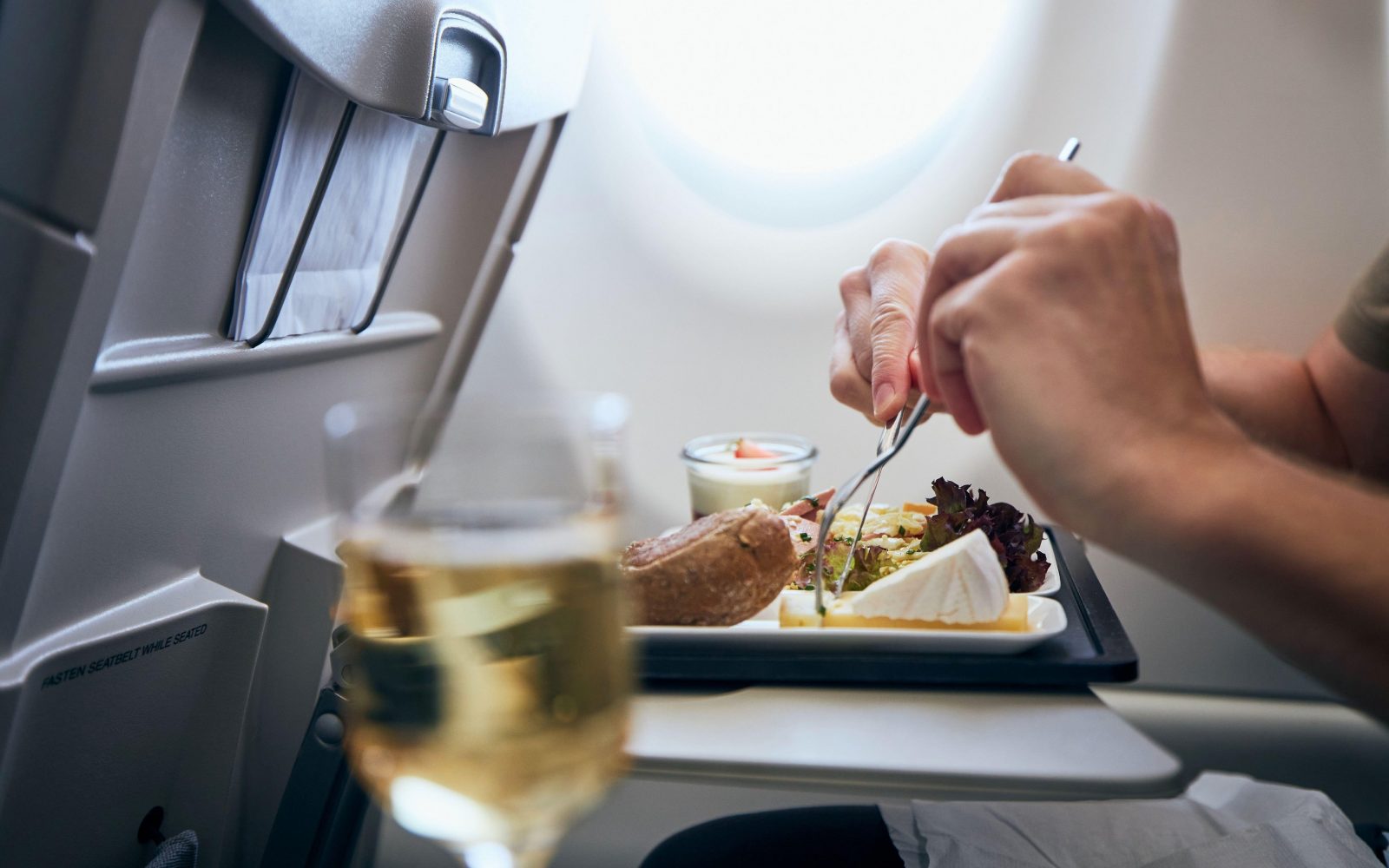 Fotografija prikazuje potnika, ki uživa obrok na letalu. Na pladnju je več jedi, vključno s kruhom, solato in sirom, ob strani pa je kozarec vina. Prizor poudarja udobje in kakovost storitve med letom, kar je pomembno za potnike na dolgih letih. Ta slika je idealna za članke o potovalnih izkušnjah, letalskih storitvah, nasvetih za potovanja in udobju na letalu.