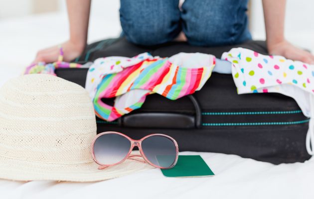 Oseba, ki pakira kovček z barvitimi oblačili, slamnikom, sončnimi očali in potnim listom..