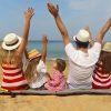 Čudovita fotografija srečne družine, ki sedi na peščeni plaži ob morju. Vsi člani družine imajo dvignjene roke in nosijo poletna oblačila ter klobuke. Morje v ozadju je mirno, nebo pa jasno, kar ustvarja popolno poletno vzdušje. Ta slika popolno zajame veselje in povezanost družine med poletnimi počitnicami ter je idealna za promocijo družinskih izletov, poletnih destinacij in sprostitvenih trenutkov na plaži.
