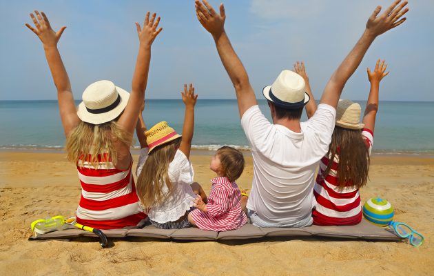 Čudovita fotografija srečne družine, ki sedi na peščeni plaži ob morju. Vsi člani družine imajo dvignjene roke in nosijo poletna oblačila ter klobuke. Morje v ozadju je mirno, nebo pa jasno, kar ustvarja popolno poletno vzdušje. Ta slika popolno zajame veselje in povezanost družine med poletnimi počitnicami ter je idealna za promocijo družinskih izletov, poletnih destinacij in sprostitvenih trenutkov na plaži.