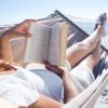 Ženska v beli čipkasti obleki leži v viseči mreži na plaži in bere knjigo. V ozadju je morje in modro nebo. Fotografija izraža sproščenost in mir na sončnem dnevu.