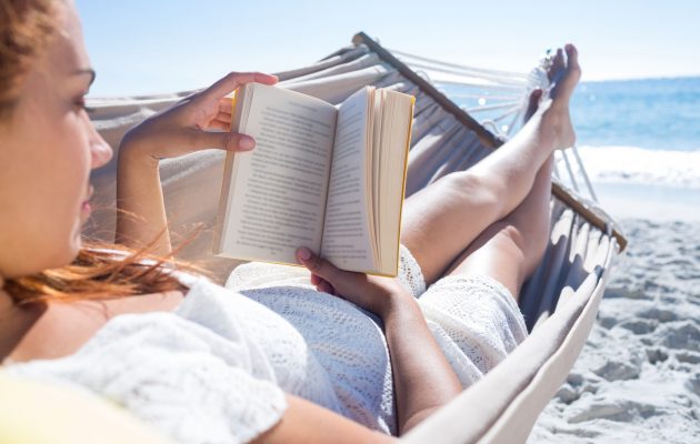 Ženska v beli čipkasti obleki leži v viseči mreži na plaži in bere knjigo. V ozadju je morje in modro nebo. Fotografija izraža sproščenost in mir na sončnem dnevu.