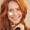 Na sliki je ženska z rdečimi, valovitimi lasmi in veselim nasmehom, z nanosom sončne kreme na licu, kar simbolizira zaščito pred soncem in nego kože. Ta slika je idealna za teme, povezane s poletno nego kože, prednostmi sončne kreme, lepotnimi rutinami in zdravo kožo. Poudarja pomen uporabe sončne kreme za zaščito pred UV žarki, hkrati pa promovira vesel, pozitiven pristop k vsakodnevni negi kože. Primerno za vsebine o dermatologiji, izdelkih za nego kože ali nasvetih za zdravje poleti.