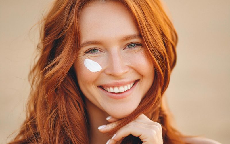 Na sliki je ženska z rdečimi, valovitimi lasmi in veselim nasmehom, z nanosom sončne kreme na licu, kar simbolizira zaščito pred soncem in nego kože. Ta slika je idealna za teme, povezane s poletno nego kože, prednostmi sončne kreme, lepotnimi rutinami in zdravo kožo. Poudarja pomen uporabe sončne kreme za zaščito pred UV žarki, hkrati pa promovira vesel, pozitiven pristop k vsakodnevni negi kože. Primerno za vsebine o dermatologiji, izdelkih za nego kože ali nasvetih za zdravje poleti.