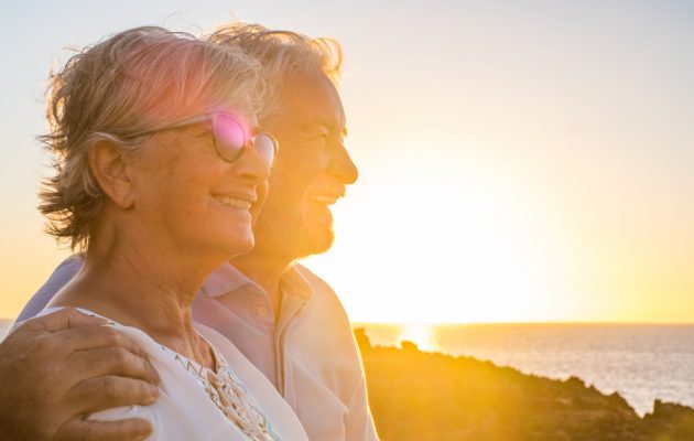 Starejši par uživa ob sončnem zahodu na plaži, nasmejani in objemajoči se, izražajoč srečo, ljubezen in sprostitev. V ozadju se razprostira morje in topla svetloba poletnega večera, kar ustvarja romantičen in miren prizor. Par nosi sproščena poletna oblačila, eden izmed njiju ima na sebi sončna očala, ki odsevajo sončne žarke. Ta slika ponazarja srečne trenutke in uživanje prostega časa v naravi med počitnicami.