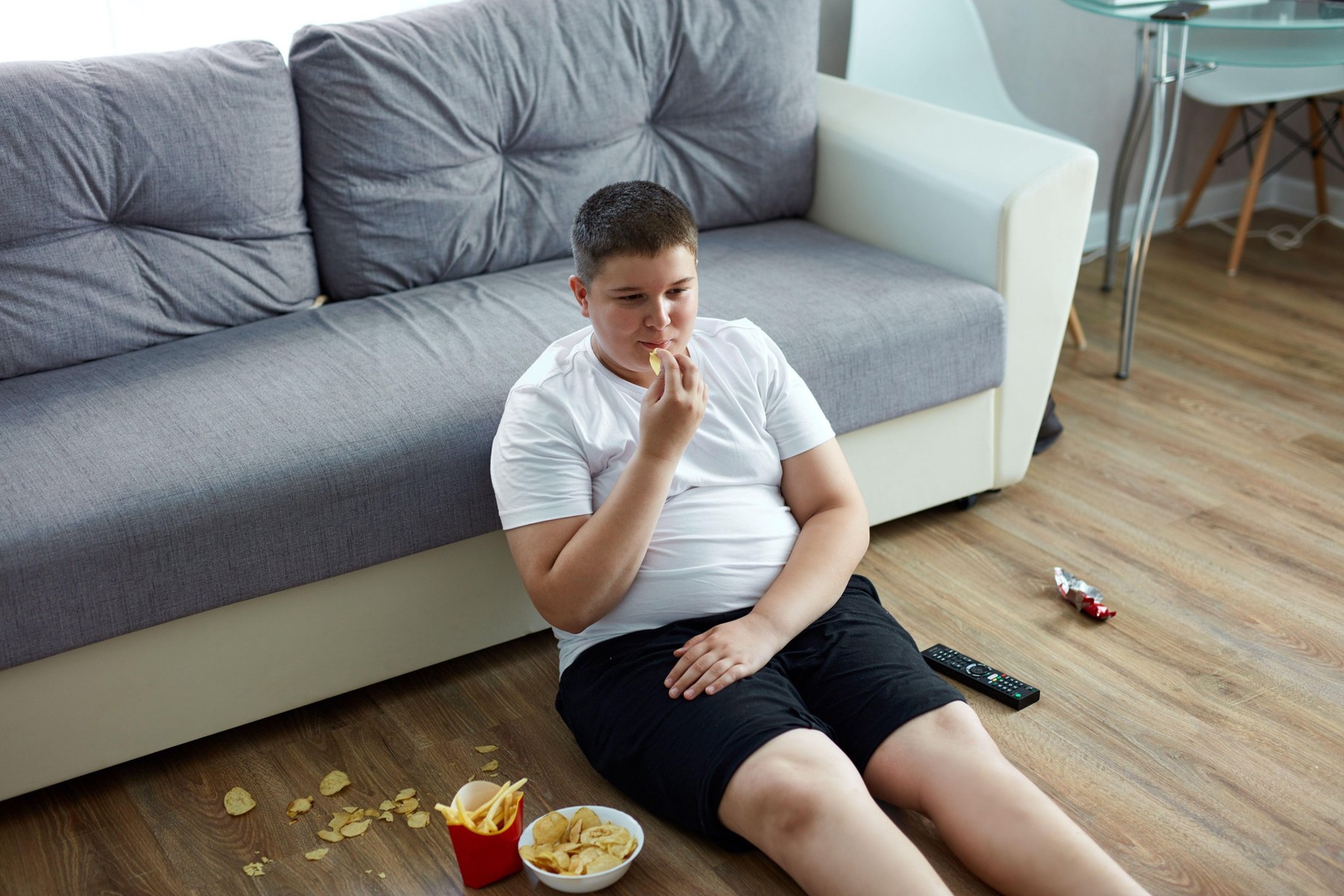 Prekomerno težek deček sedi na tleh v dnevni sobi, okoli njega je nezdrava hrana.