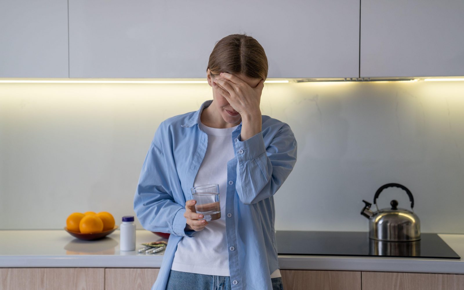 Ženska stoji v kuhinji in se drži za glavo zaradi glavobola, ki je posledica dehidracije. Je v bolečinah in slabe volje, v roki pa drži kozarec vode.