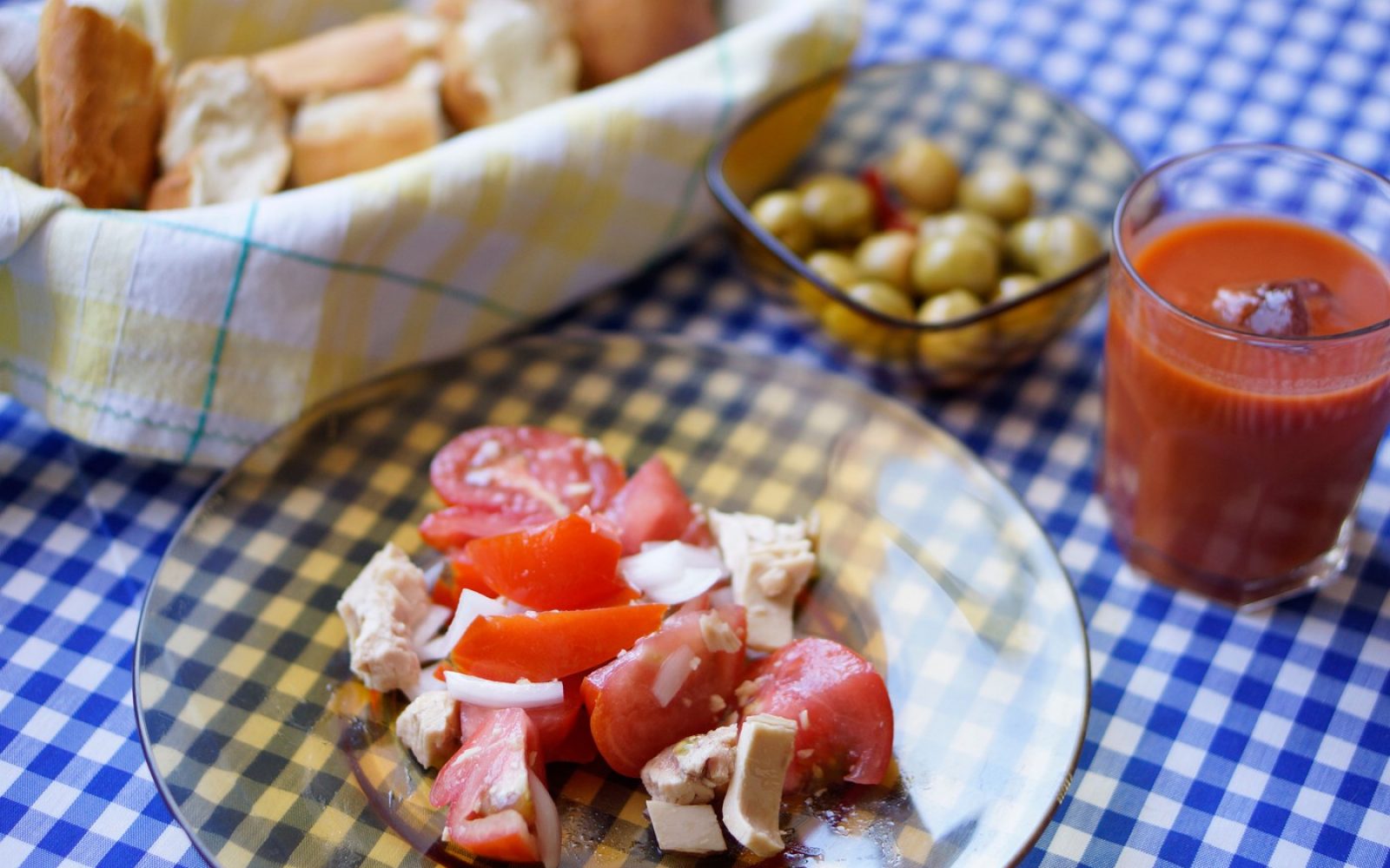 Mediteranska dieta: krožnik s paradižnikom, sirom in čebulo, ob njem kruh in skleda z olivami na karirastem prtu.