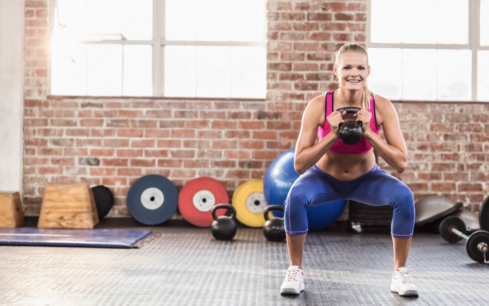 Ženska v fitnesu, polnem pisanih uteži, dela počep in pri tem drži kettlebell utež pred prsmi.