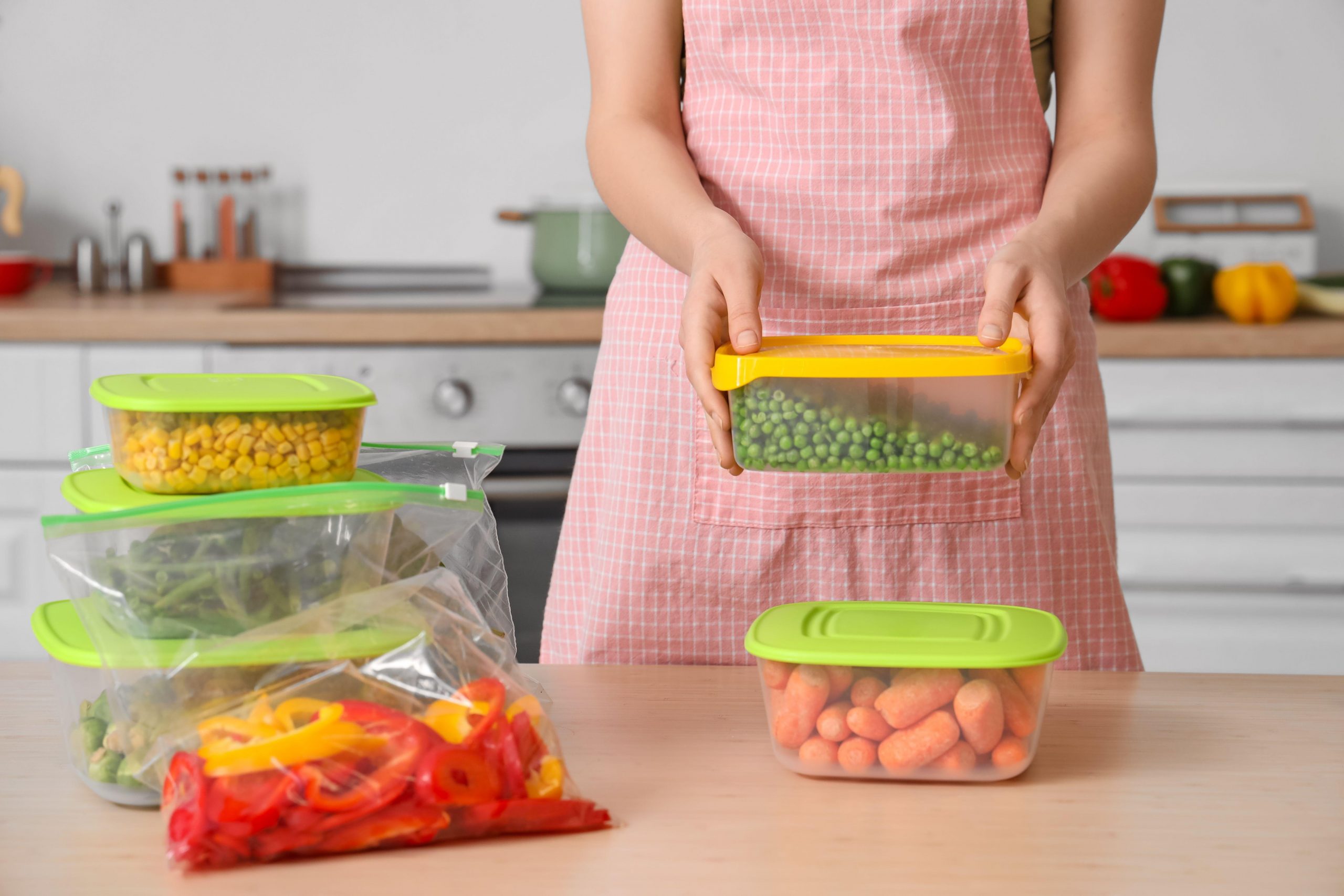 Ženska v predpasniku v roki drži posodo z grahom za zamrzovanje, na mizi pred njo pa so v neprodušno zaprtih posodah za zamrzovanje in ustreznih vrečkah še paprike, korenje, grah, ohrovt, fižol in koruza.