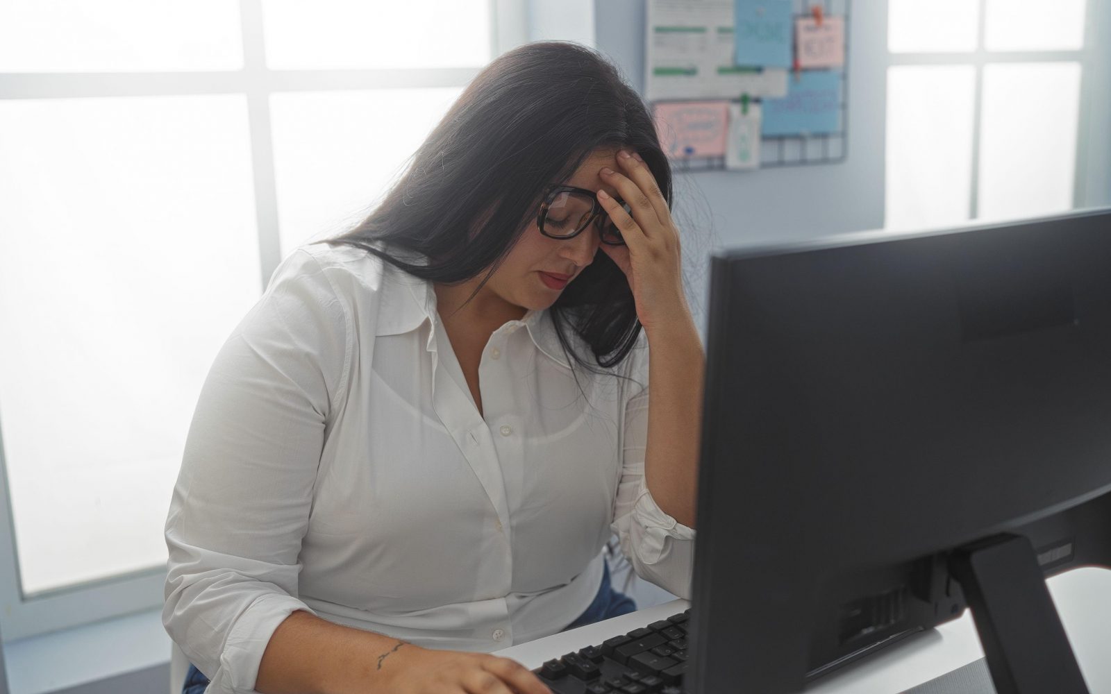 Pisarniška delavka, ki sedi za računalnikom in se drži za glavo, simbolizira stres, premalo gibanja in naraščanje telesne teže zaradi takega življenjskega sloga.