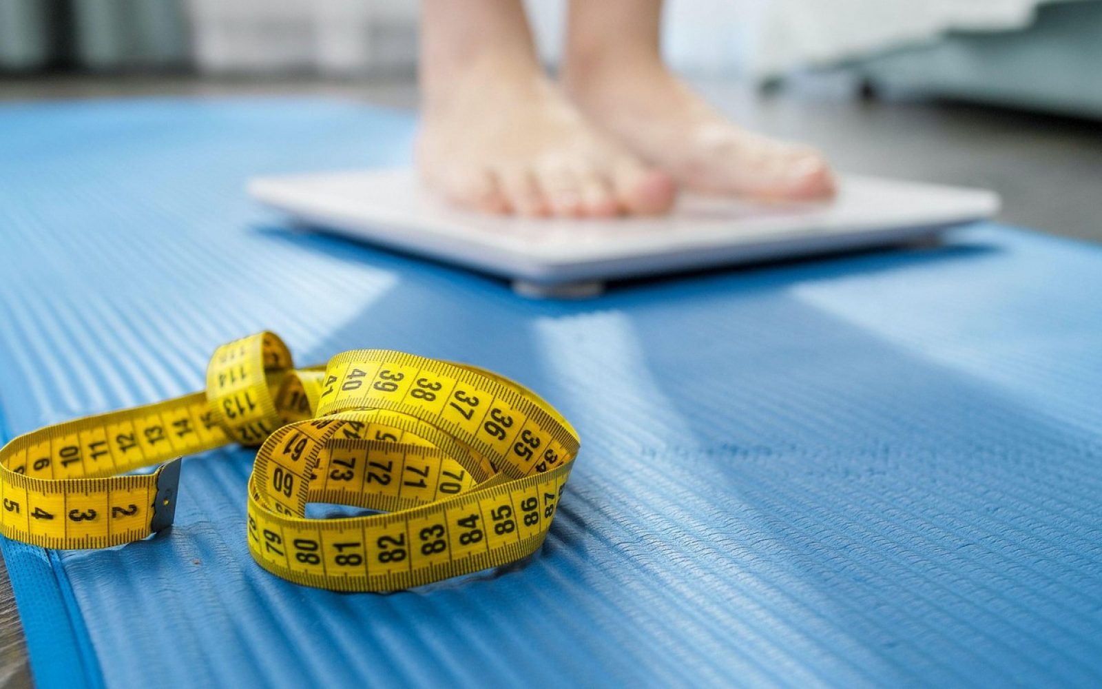 Oseba stoji na tehtnici, ob njej pa je meter – simbol hujšanja in diete 1200 kalorij na dan.