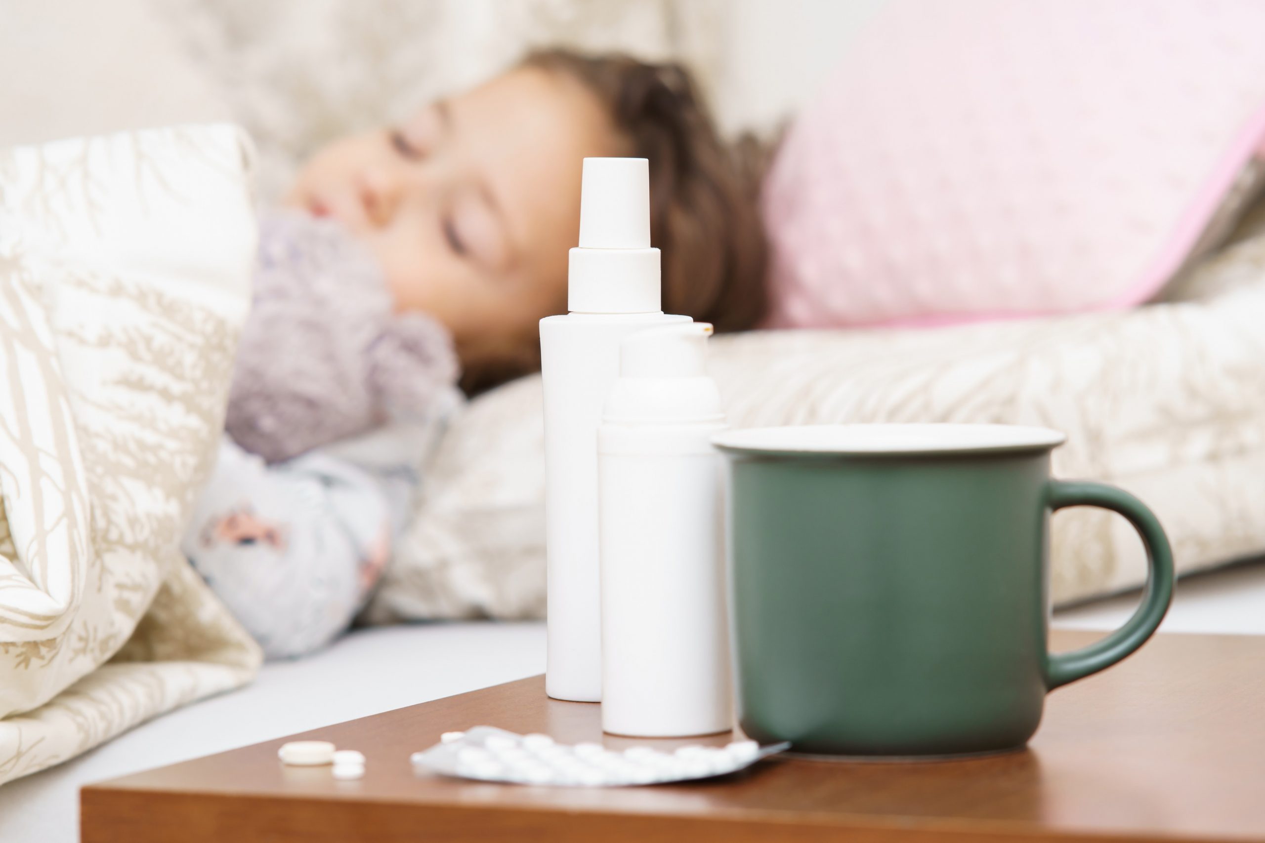 Mala bolna in prehlajena punčka spi v postelji, na nočni omarici ima sprej za nos, tablete in šalico čaja.