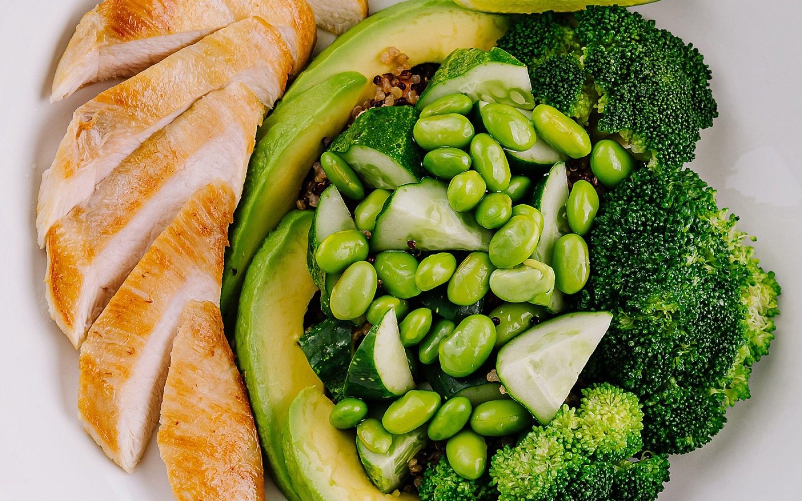 Uravnotežen obrok na krožniku z rezinami pečenega piščanca, avokadom, brokolijem, edamamom in limeto.