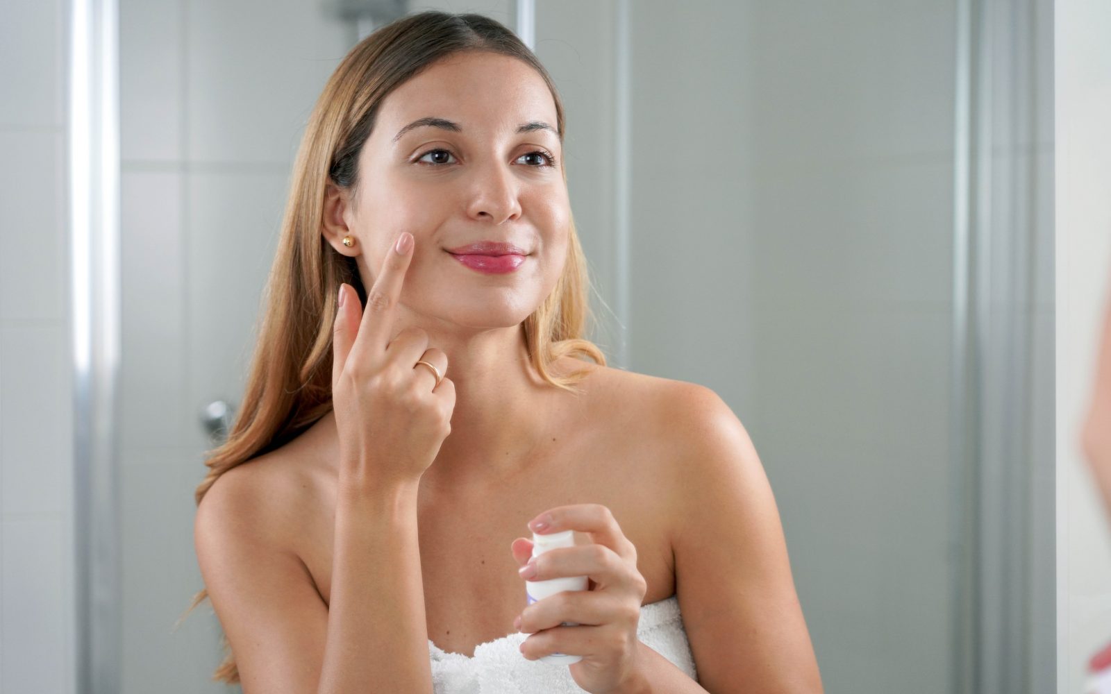 Mlada ženska si v kopalnici na obraz nanaša vlažilno kremo - večerna rutina za sijočo in prožno kožo