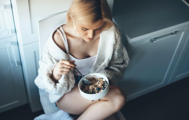 Mlada ženska v udobni pleteni jopi uživa v skledi granole z jogurtom in sadjem na tleh v kuhinji.