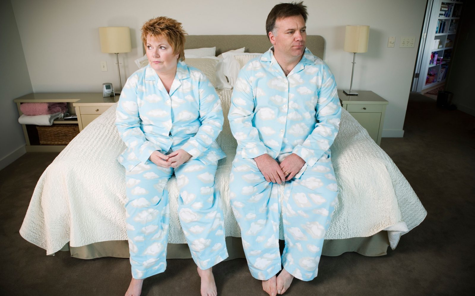 Moški in ženska srednjih let v ujemajočih se pižamah sedita na robu postelje – ponazoritev naraščanja telesne teže in nezdravega življenjskega sloga v Sloveniji.