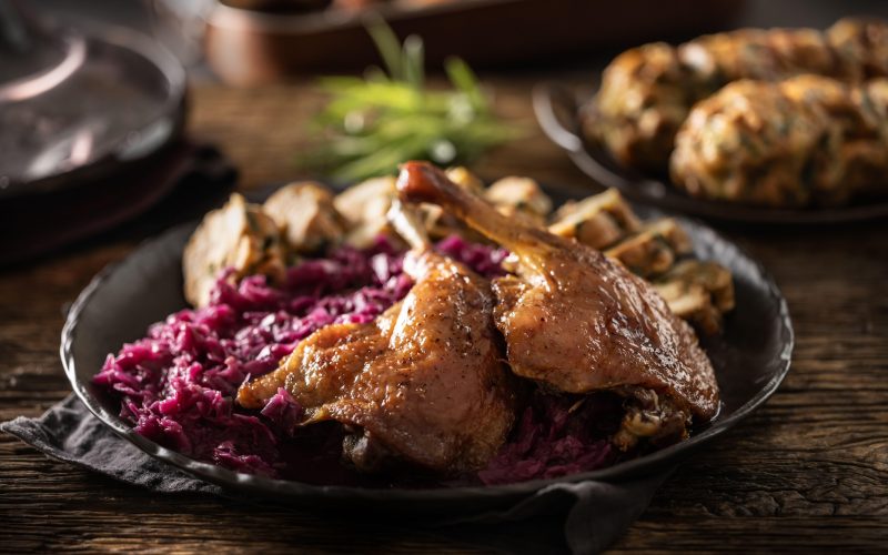 Slika mize ob martinovi pojedini - pečena goska, dušeno rdeče zelje, mlinci in kruhovi cmoki na temnem krožniku, v ozadju vino in druge jesenske priloge