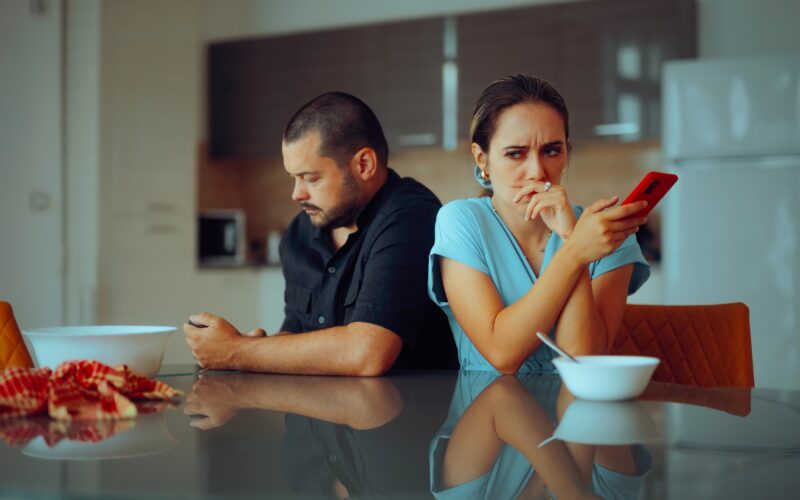Odtujeni par sedi za mizo, obrnjena sta drug proti drugemu s hrbtom, vsak ima v rokah svoj telefon