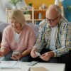 Starejši par sedi na kavču, nekaj skupaj delata, ona ima v rokah telefon, on listek