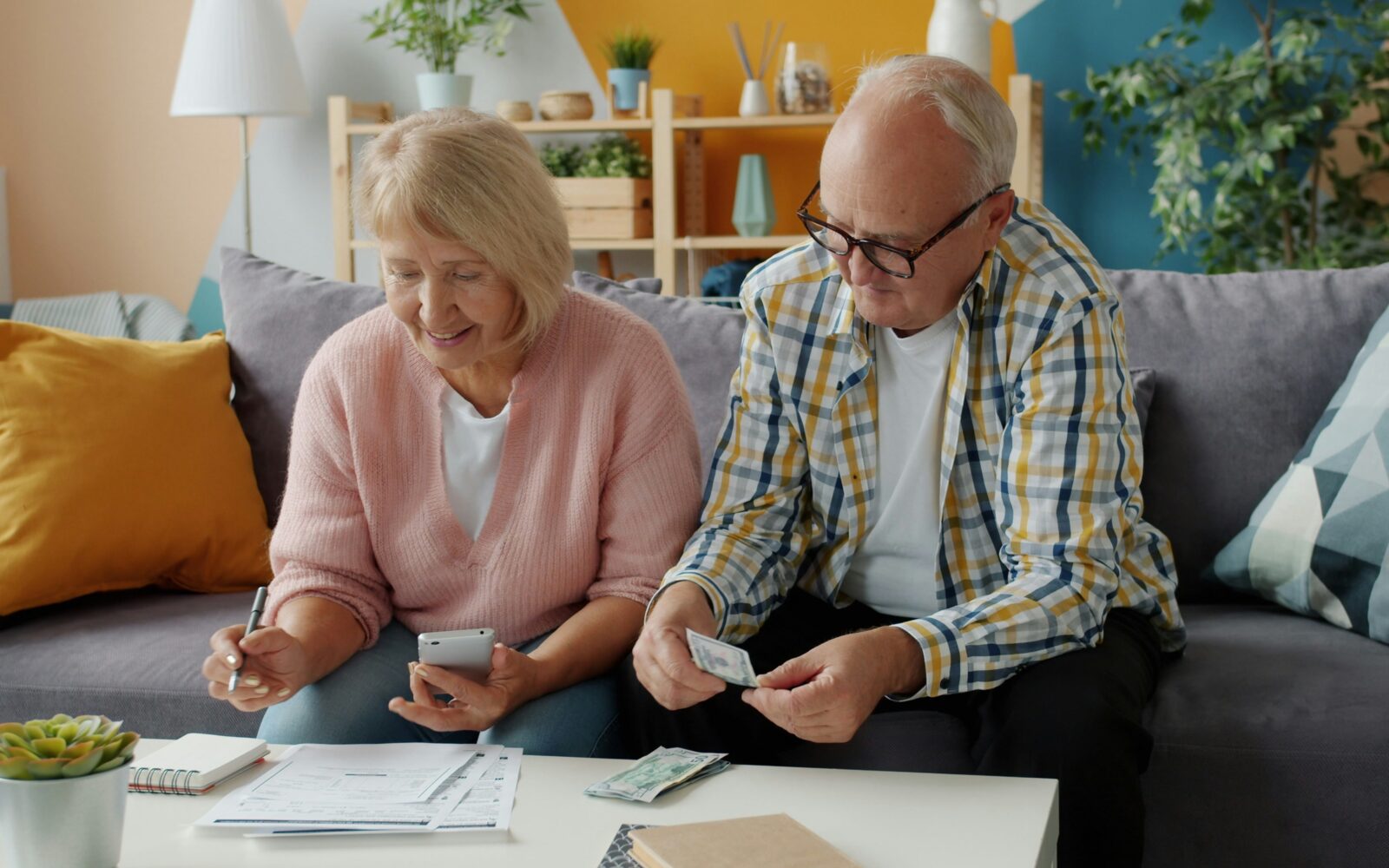 Starejši par sedi na kavču, nekaj skupaj delata, ona ima v rokah telefon, on listek