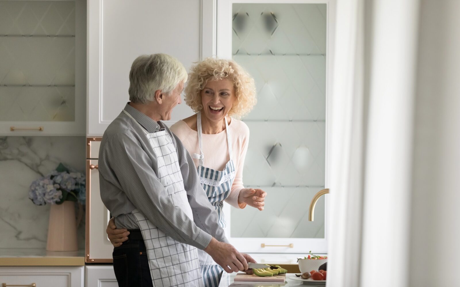 Starejši par je nasmejan v kuhinji, oba imata predpasnike in se smejita