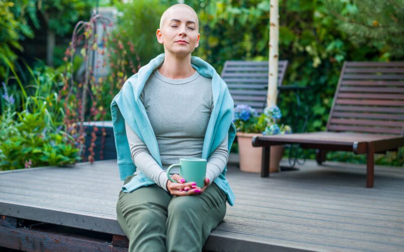 Ženska z rakavo boleznijo je izgubila lase. Sedi na podestu pred hišo, v rokah drži skodelico.