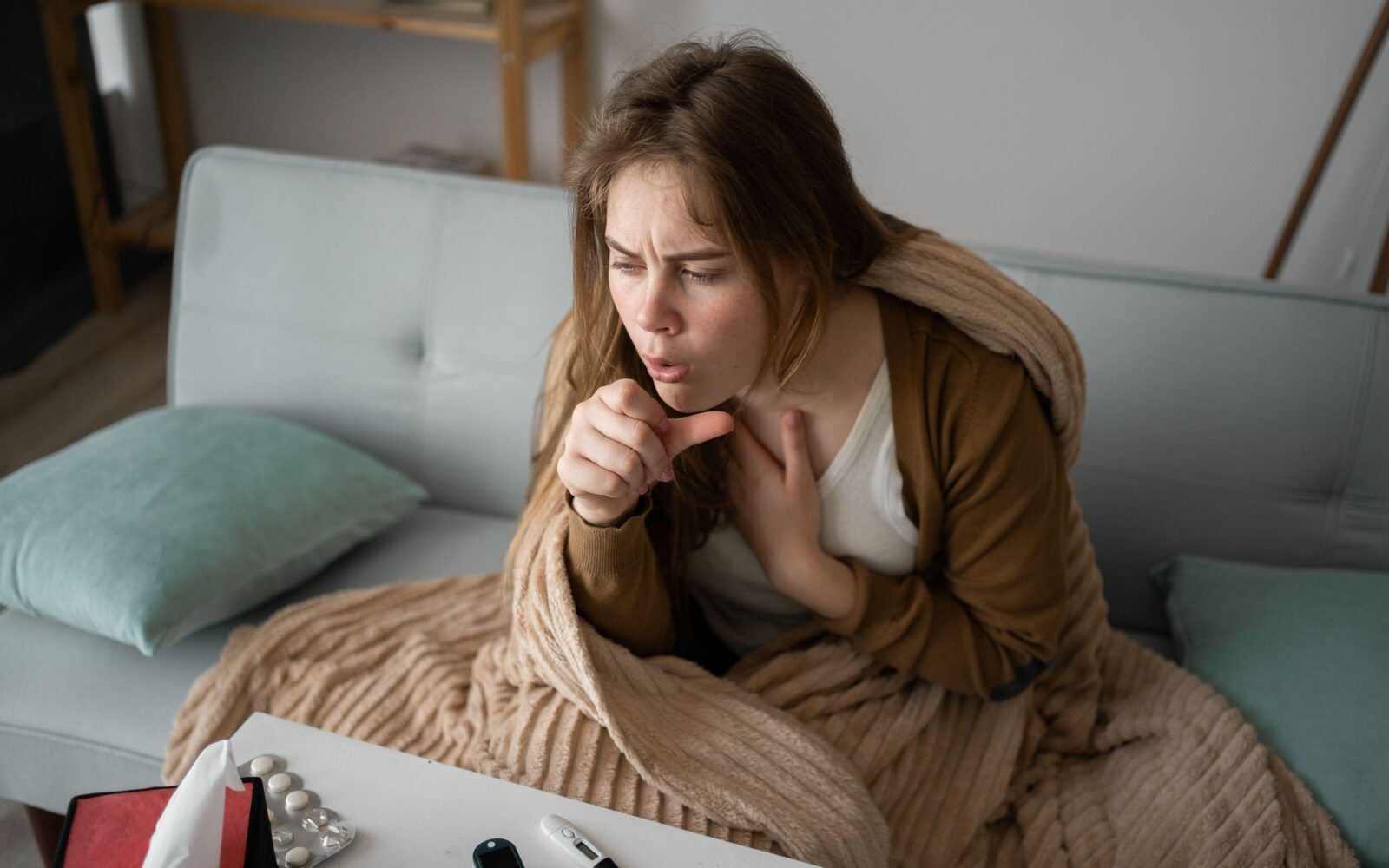 Mlada bolna ženska sedi na kavču zavita v odejo in kašlja.