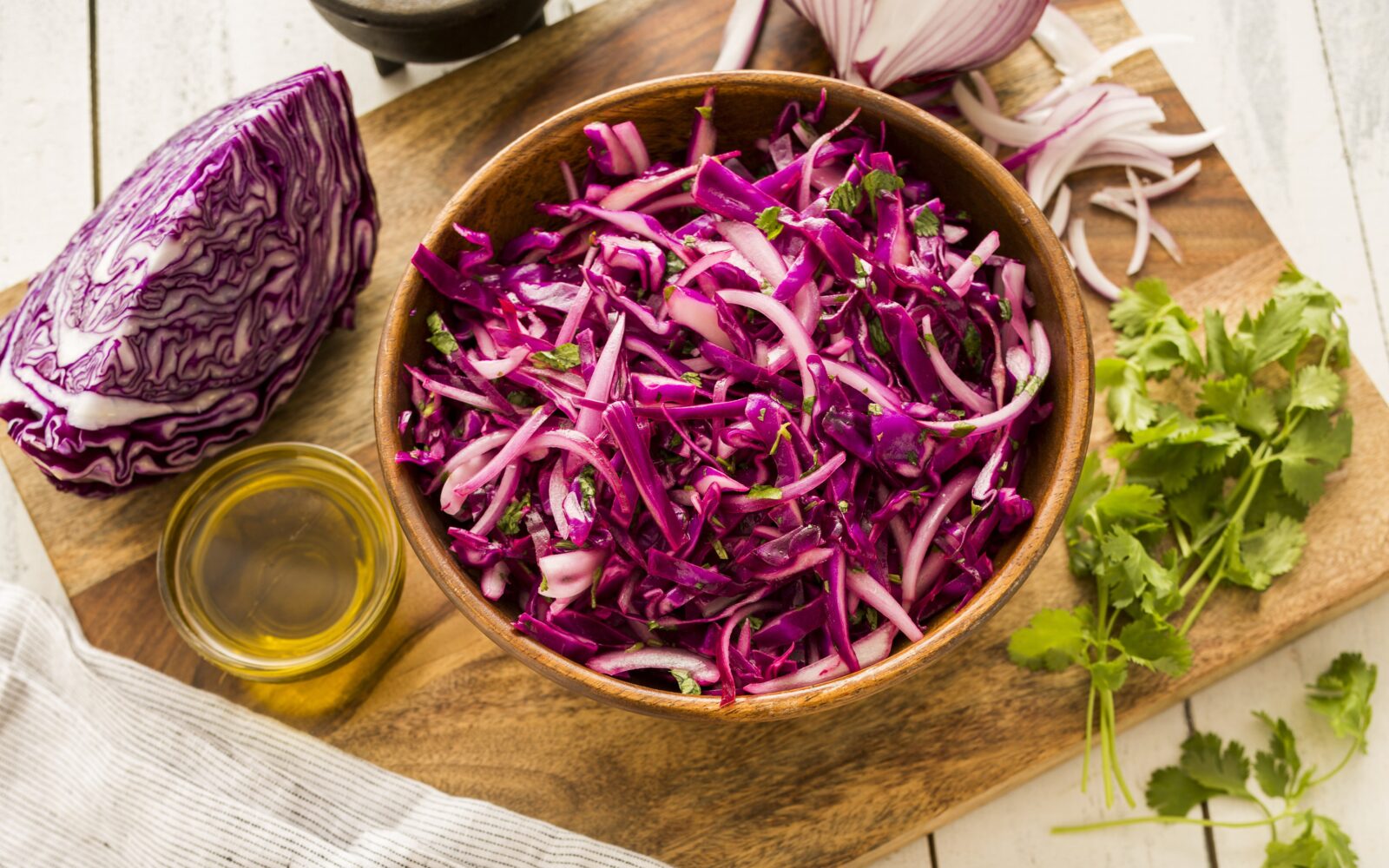 Sveža solata iz rdečega zelja v leseni skledi, ob njej pa olje, zelišča in narezane sestavine.