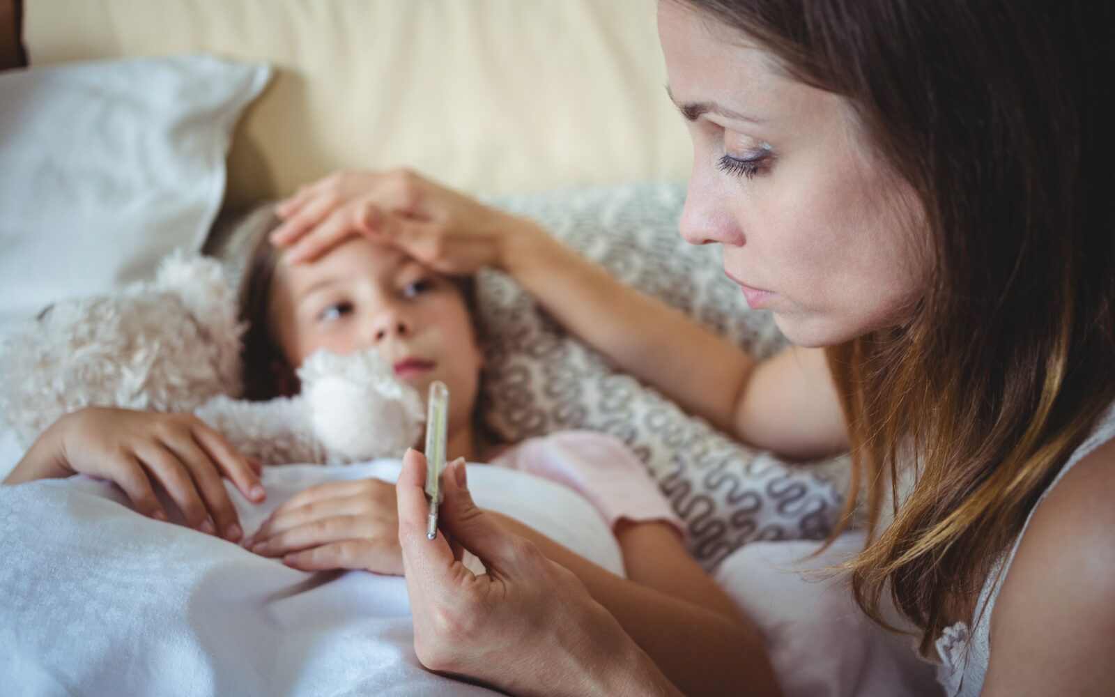 Bolna deklica leži, mati ji je izmerila temperaturo, v rokah drži termometer in jo drži za čelo