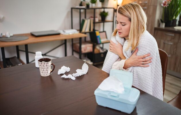 Bolna ženska sedi za mizo, pred sabo ima skodelico in posmrkane robčke
