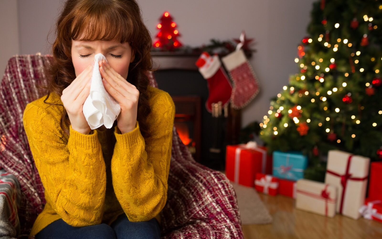 Ženska v rumenem puloverju sedi na kavču, in si briše izcedek iz nosu. Za njo je okrašena novoletna smrečica, pod katero so darila