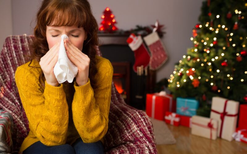 Ženska v rumenem puloverju sedi na kavču, in si briše izcedek iz nosu. Za njo je okrašena novoletna smrečica, pod katero so darila