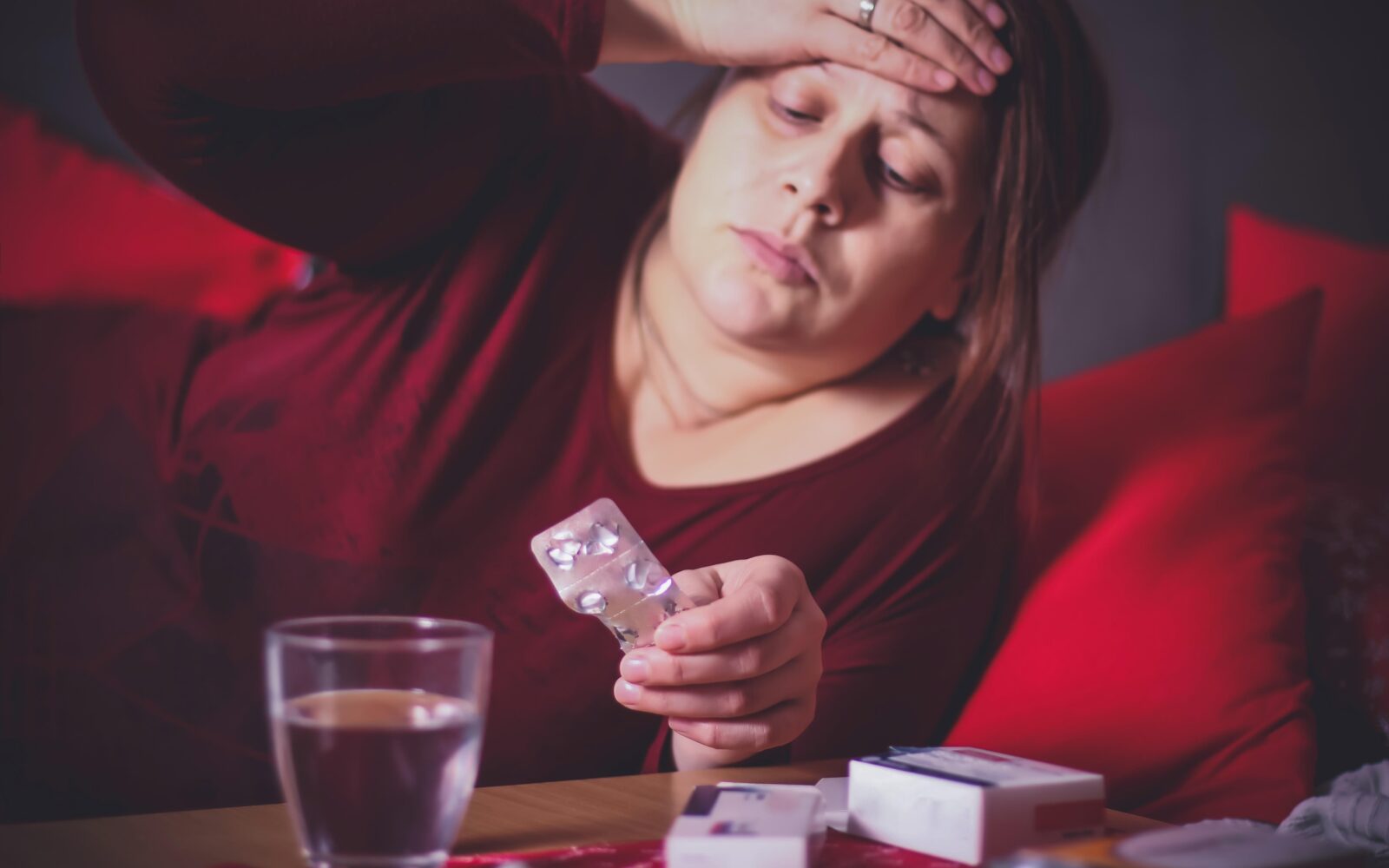 Bolna ženska se drži za čelo, preverja ali ima povišano telesno temperaturo, pred sabo ima kozarec vode in zdravila