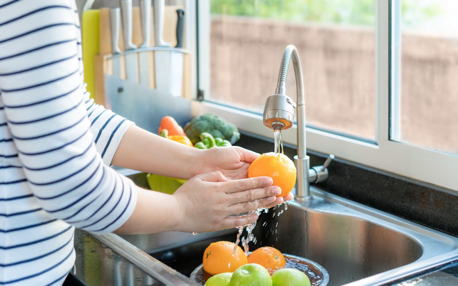 Ženska pod tekočo vodo pere pomaranče na kuhinjskem pultu.