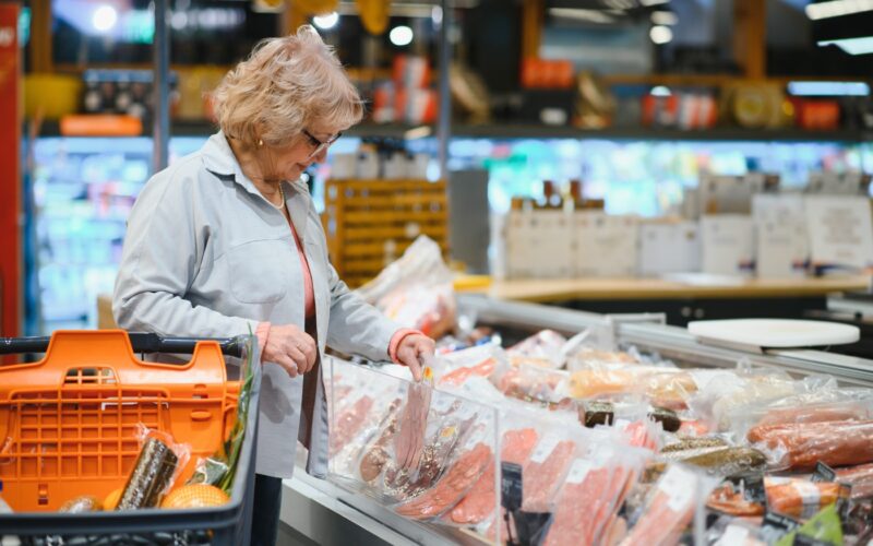 Starejša ženska v trgovini izbira pakirano meso iz hladilne vitrine.