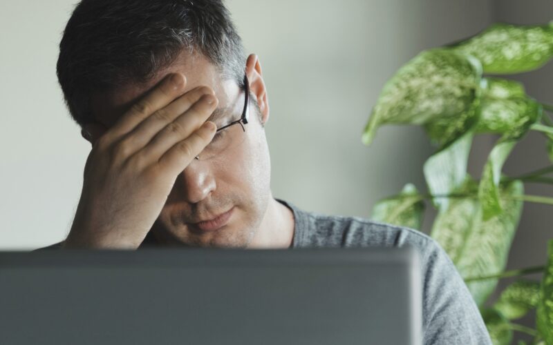 Moški z očali sedi za računalnikom, gleda v ekran in se drži za glavo. Morda je utrujen, morda ga boli glava
