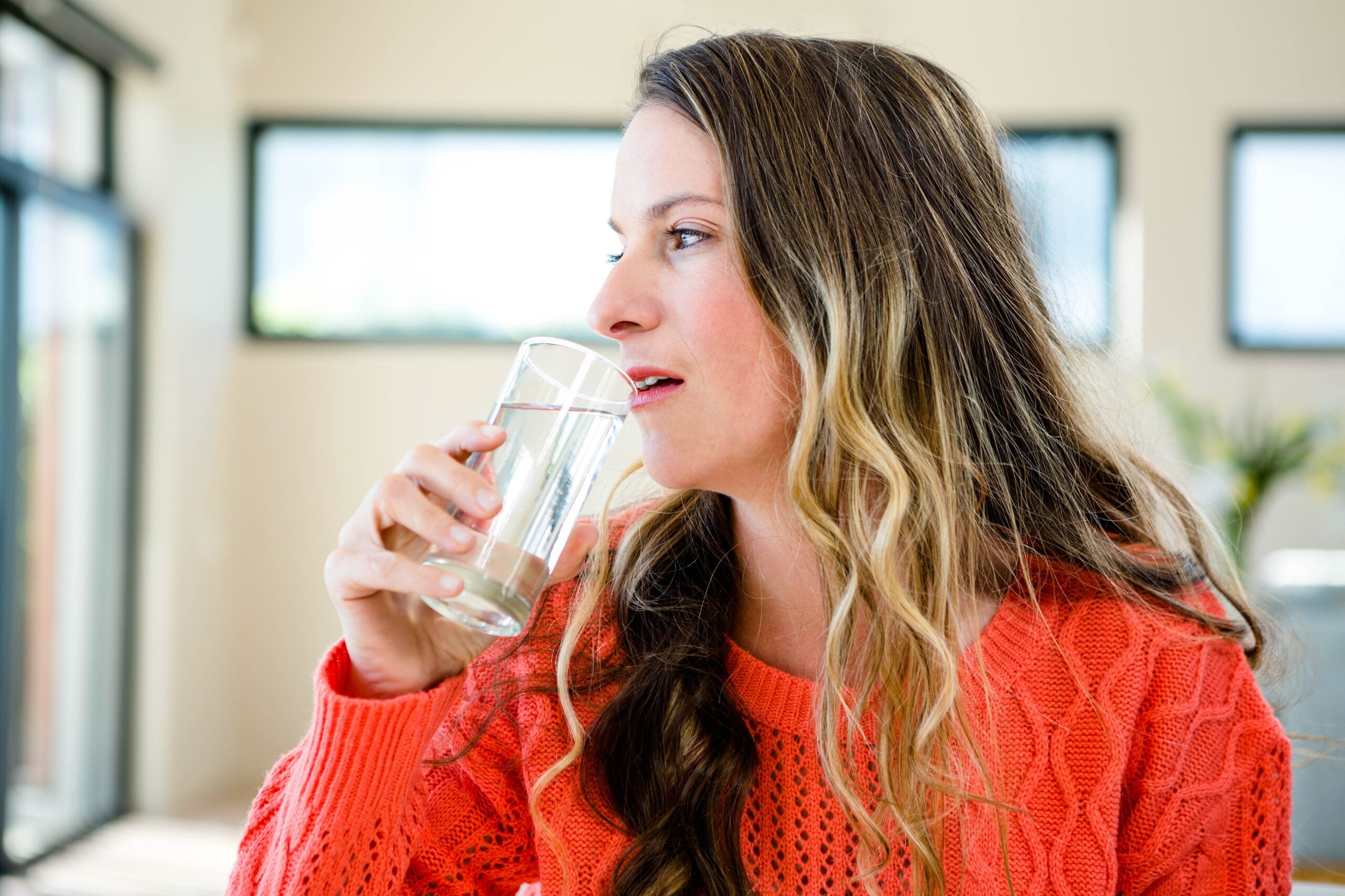 Ženska z dolgimi lasmi, oblečena v oranžni pulover pije vodo