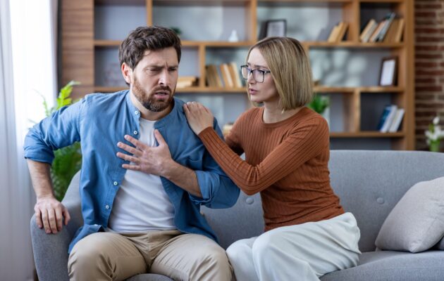 Moški in ženska sedita na kavču, moški j zadihan, občuti tiščanje v prsih, ženska ga zaskrbljeno gleda