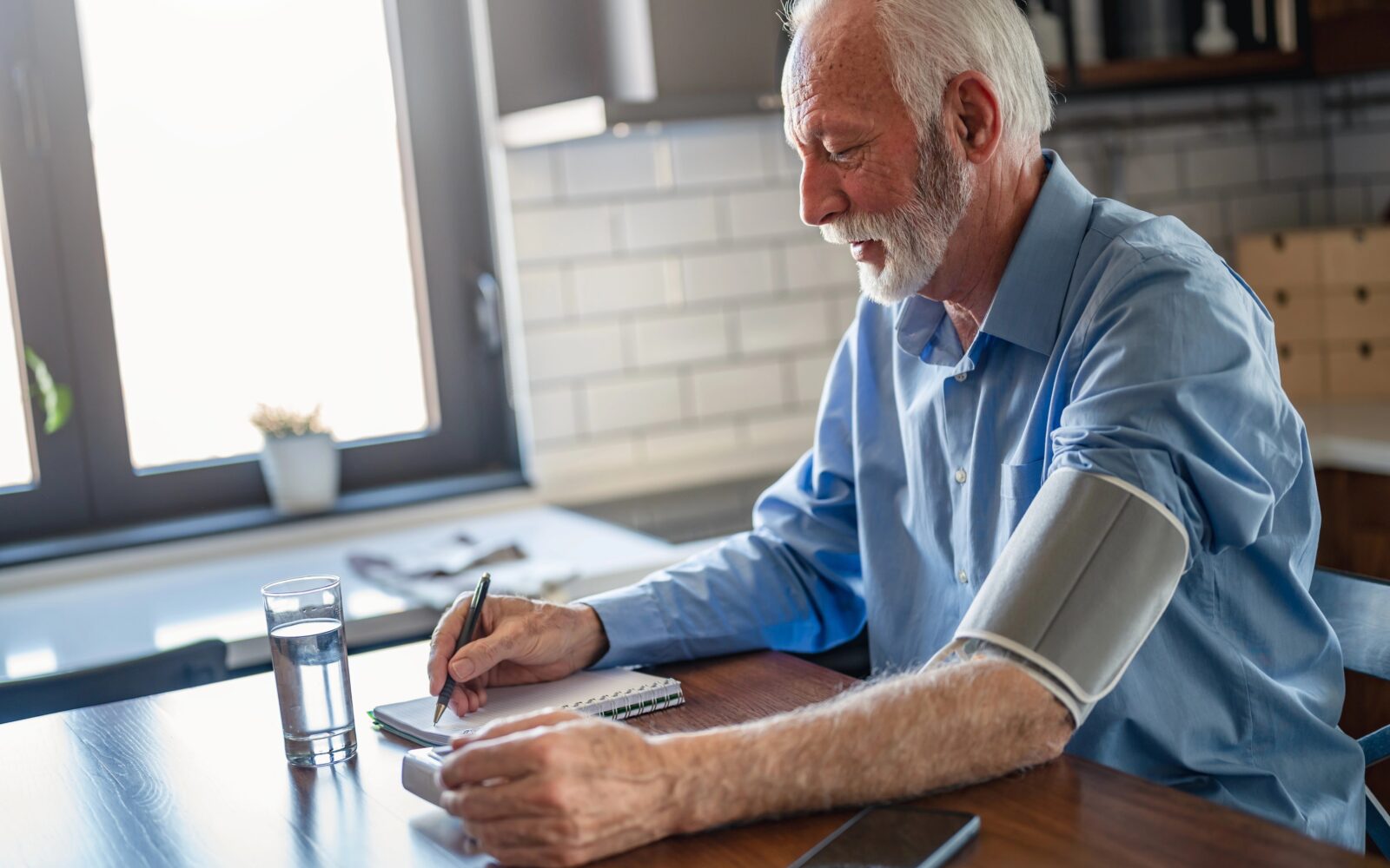 Starejšo moški si doma meri krvni tlak na levi roki, z desno pa piše izmerjene meritve