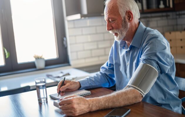 Starejšo moški si doma meri krvni tlak na levi roki, z desno pa piše izmerjene meritve