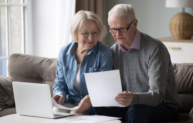 Nasmejana upokojenca sedita na kavču doma s prenosnim računalnikom in razvrščata papirje