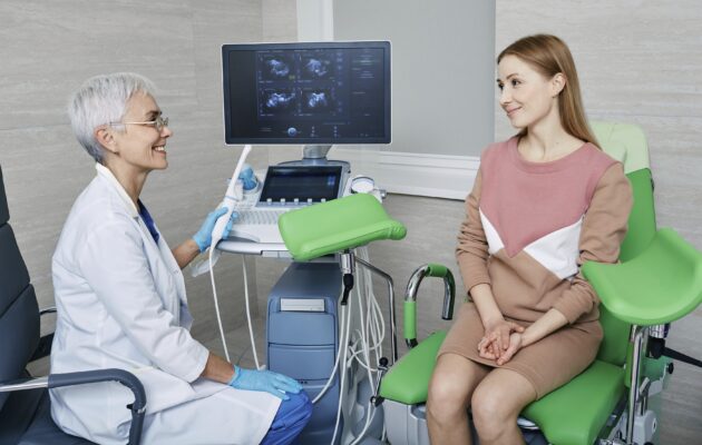 Ženska je na pregledu pri ginekologinji, sedi na zelenem ginekološkem stolu, ginekologinja pa ji nekaj razlaga