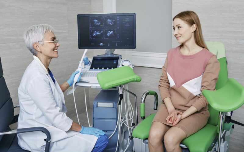 Ženska je na pregledu pri ginekologinji, sedi na zelenem ginekološkem stolu, ginekologinja pa ji nekaj razlaga