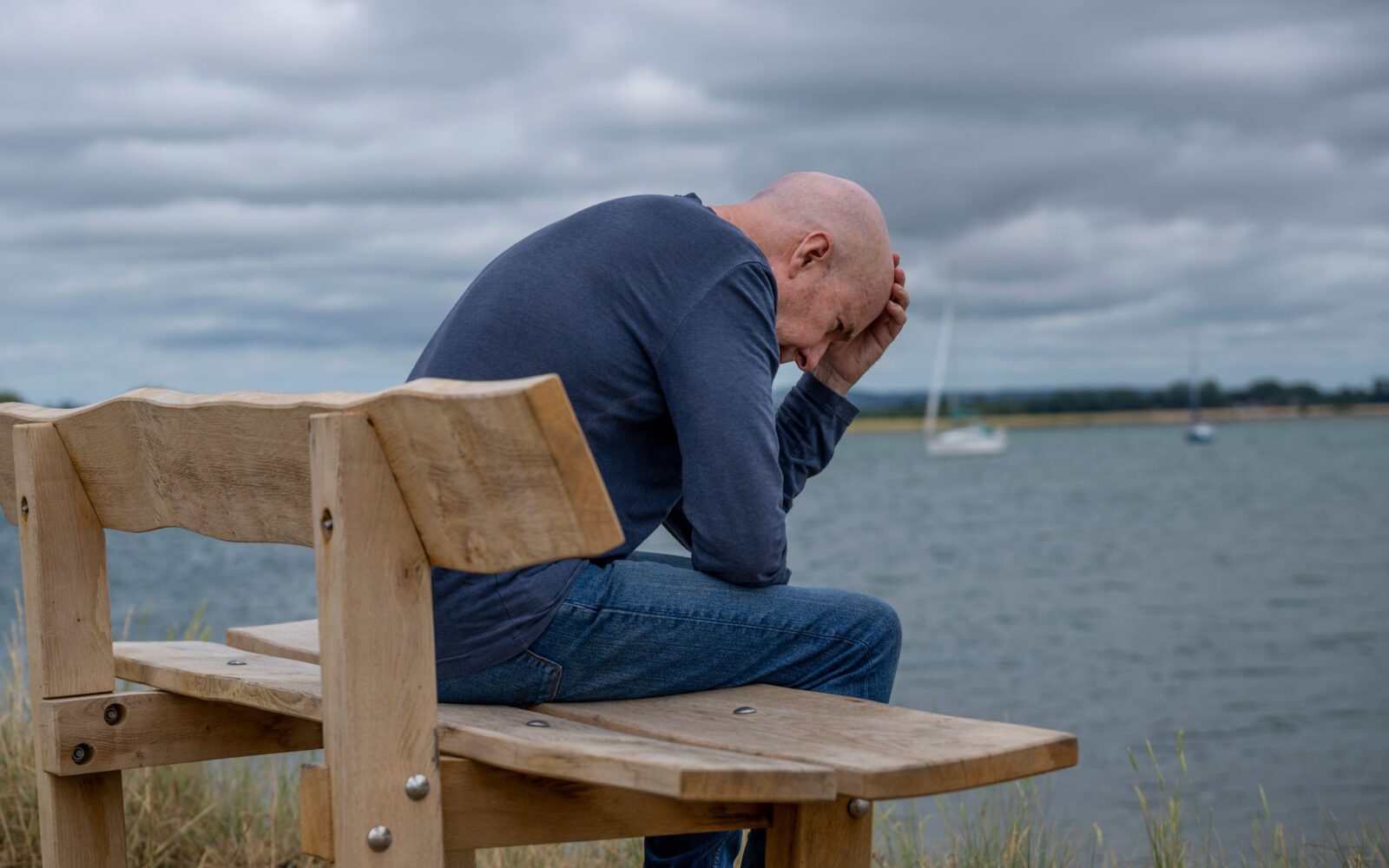 Moški na bolniški sedi ob morju, vidno izčrpan, vprašanje ostaja, ali je bivanje v tujini med bolniško dovoljeno.