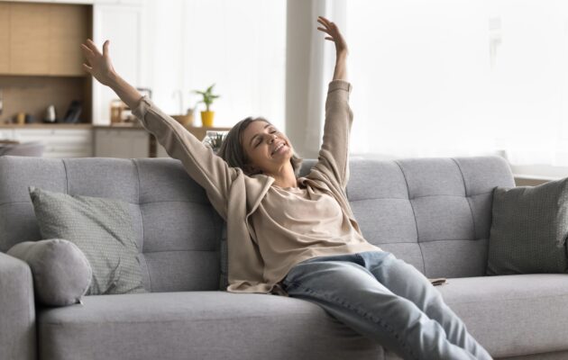 Sproščena ženska sedi na kavču, roke ima dvignjene v zrak, je vesela in nasmejana